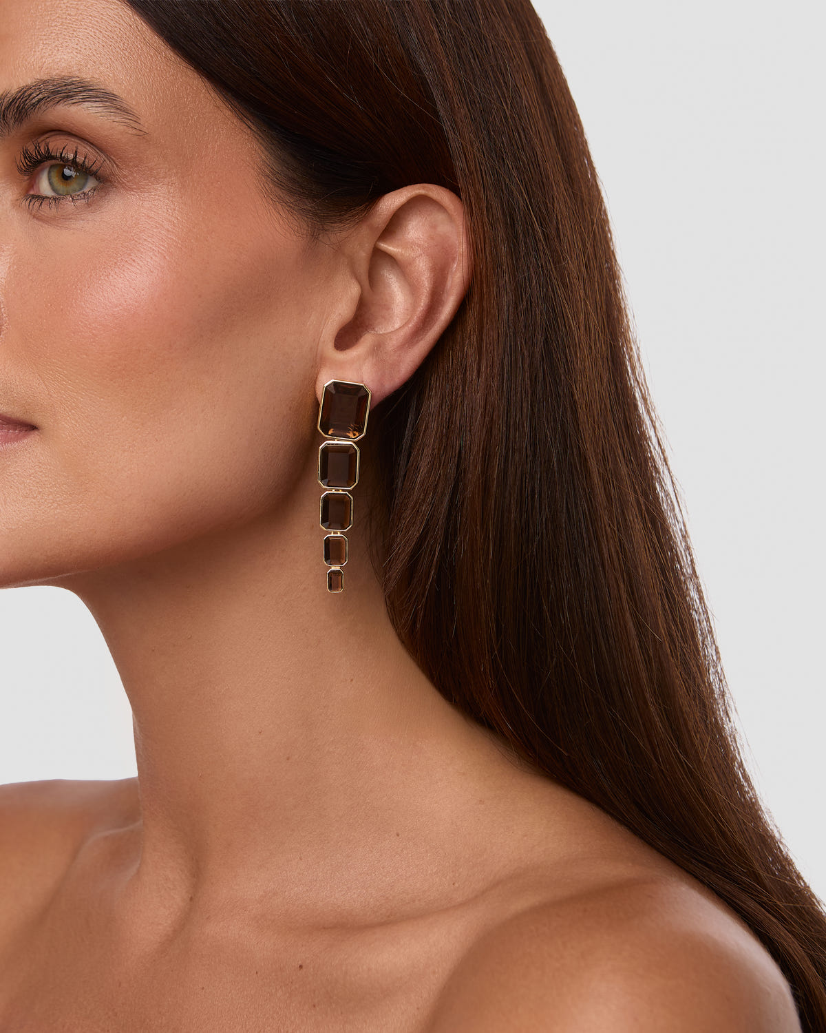 A close-up of a woman's ear wearing a gold drop earring with three stacked rectangular dark brown stones.